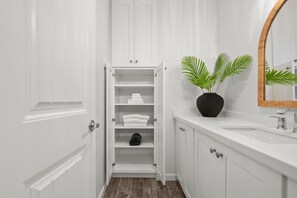 A bathroom with a white vanity, a mirror, and a shelf unit with folded towels. - 2-bedroom condo with OCEAN VIEW, A/C, KING BED, STEPS AWAY FROM SAND (Oceanside)