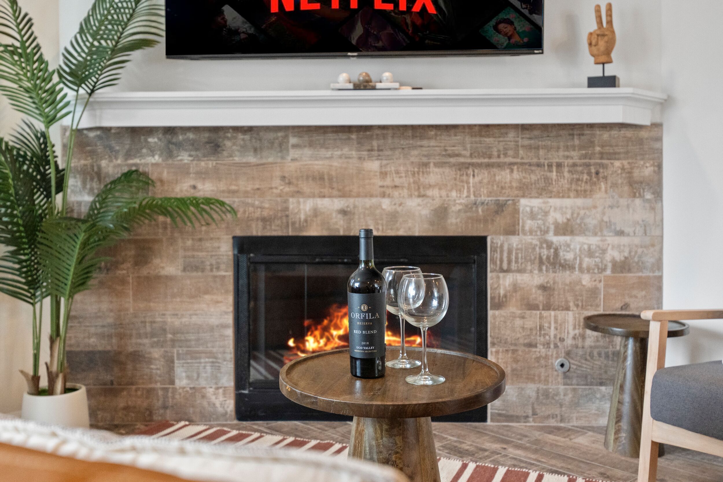 Un salon avec une cheminée, un téléviseur fixé au-dessus, une bouteille de vin et deux verres à vin sur une petite table en bois.