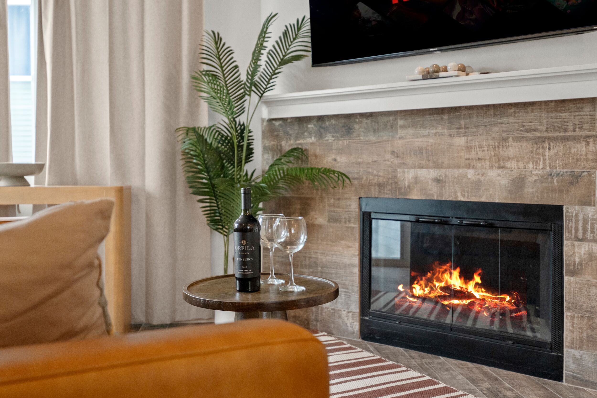 A living room with a fireplace, a sofa, a small table with a bottle and glasses, and a fern plant.