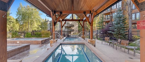 A covered outdoor swimming pool with wooden beams and a clear view of the surrounding buildings and trees.