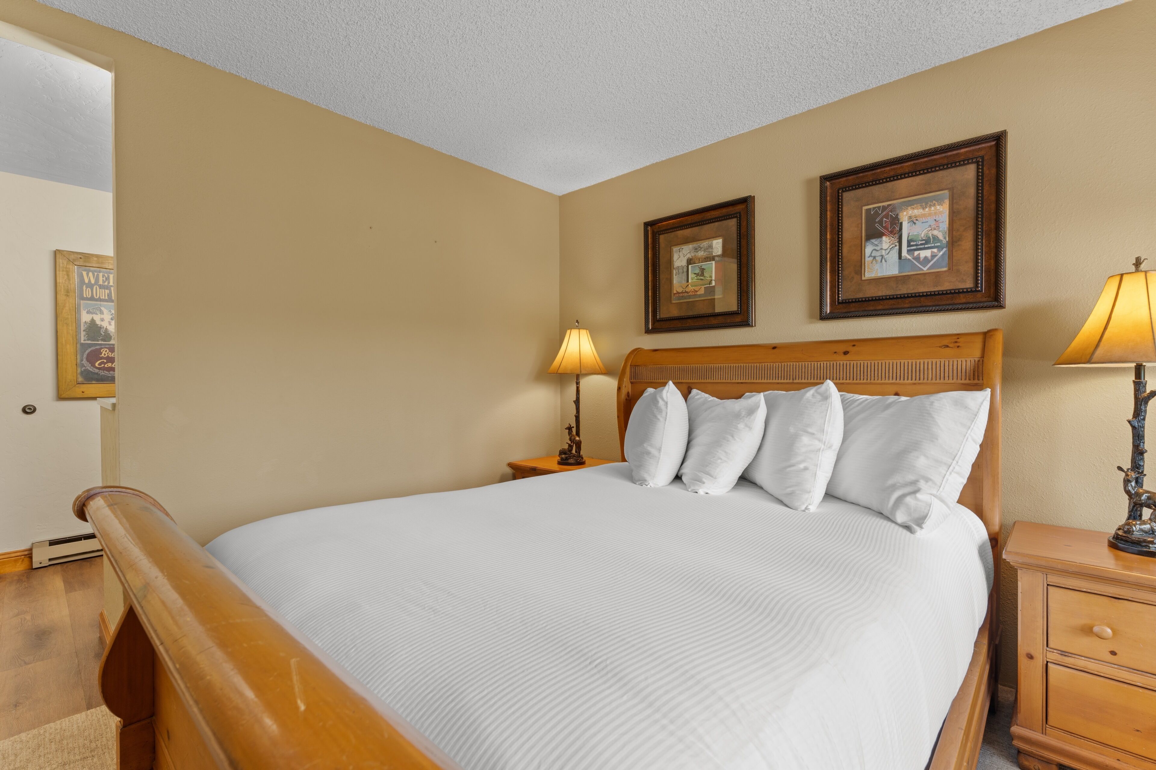 Habitación de hotel con cama de madera, ropa de cama blanca, dos cuadros en la pared, una lámpara de noche y una mesita de noche de madera.