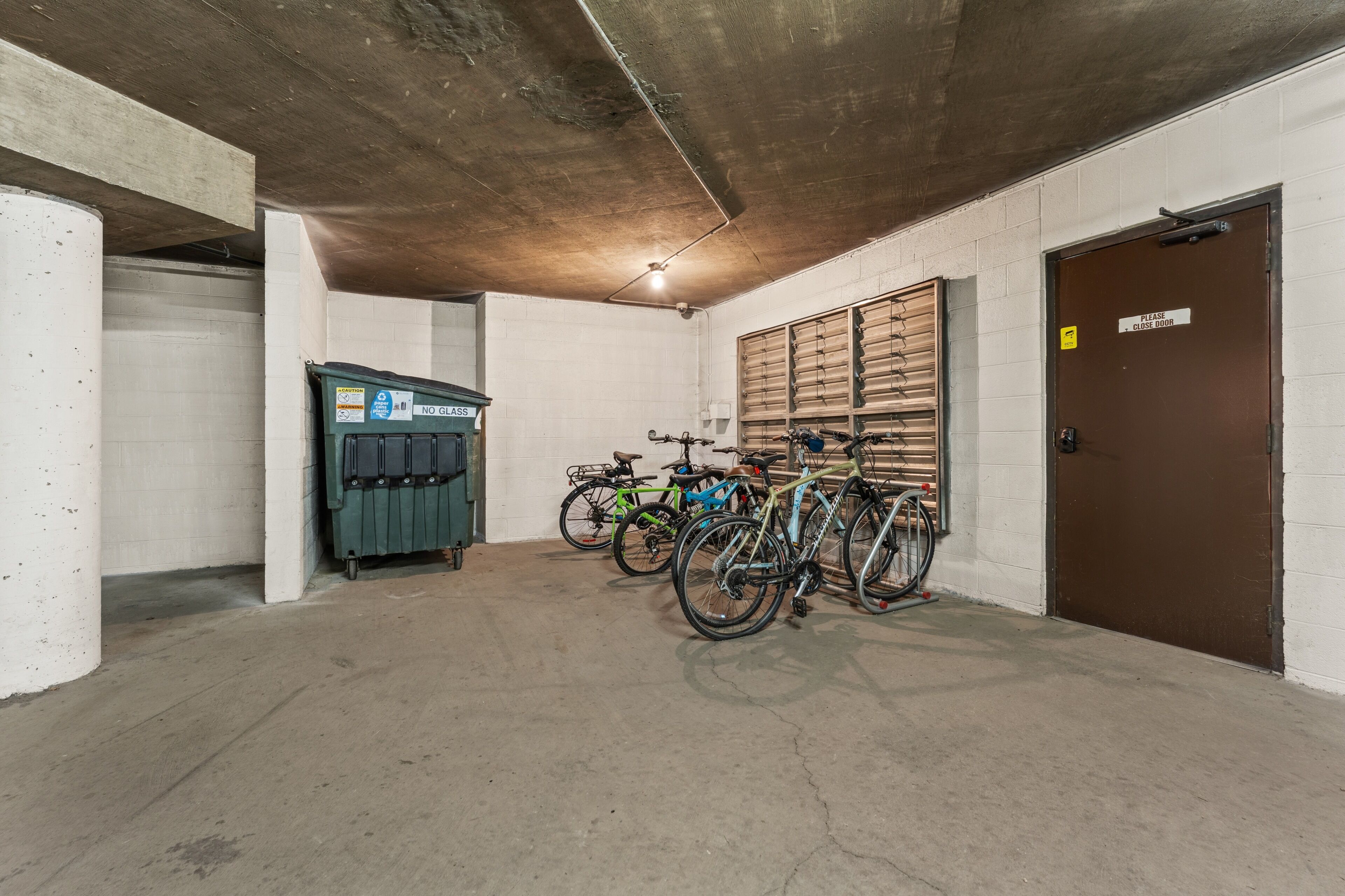 Un parking à vélos avec des bicyclettes appuyées contre le mur, une poubelle et une porte fermée avec une pancarte.