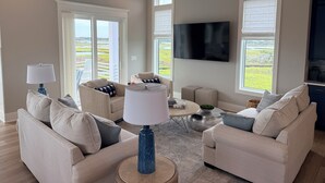 A spacious living room with a ceiling fan, large windows, a flat-screen TV, and a view of the outdoors. - Soundfront hot tub and rooftop deck (Surf City)
