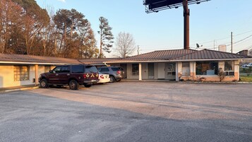 A motel with a single-story structure, a parking lot, and a row of trees in the background.
