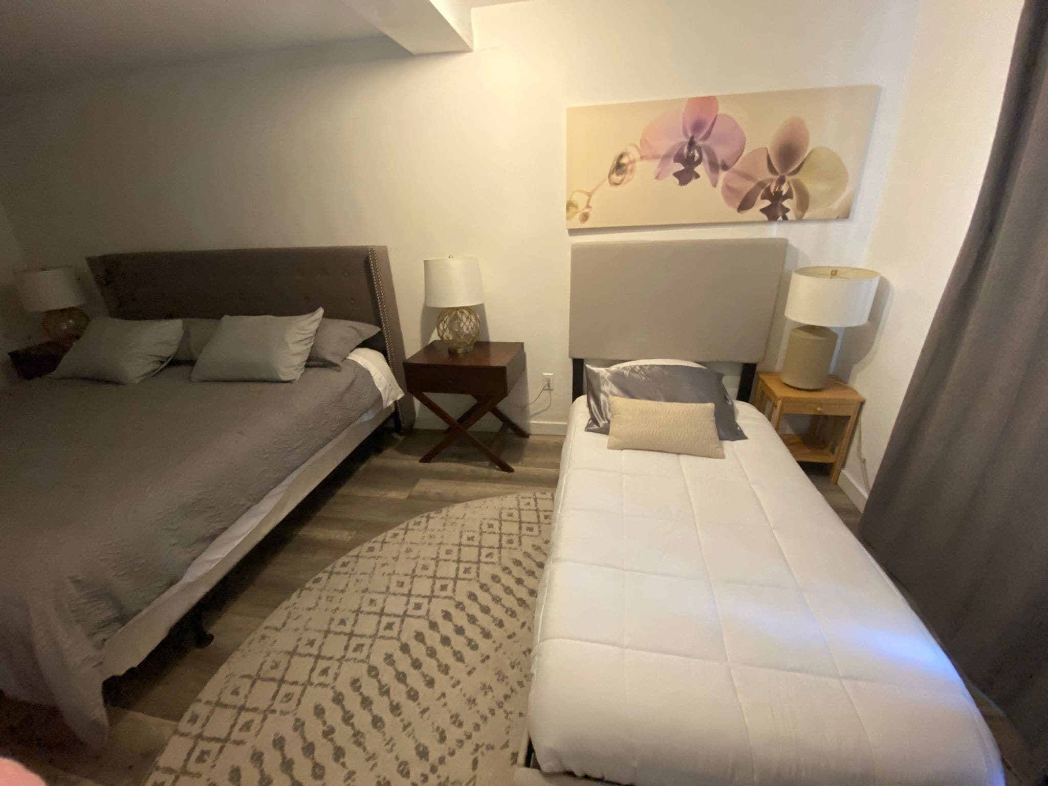 Ein Hotelzimmer mit zwei Betten, einem Nachttisch mit Lampen, einem gemusterten Teppich und einem floralen Gemälde an der Wand.
