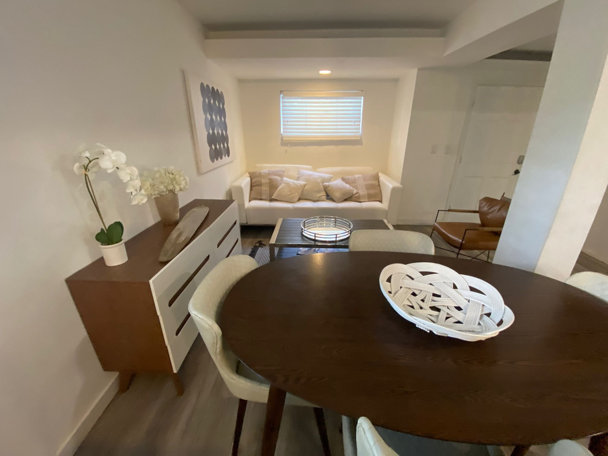 Ein Essbereich mit einem runden Tisch, Stühlen, einem weißen Dekorationsobjekt, einer Couch mit Kissen und einem Fenster mit Jalousien.