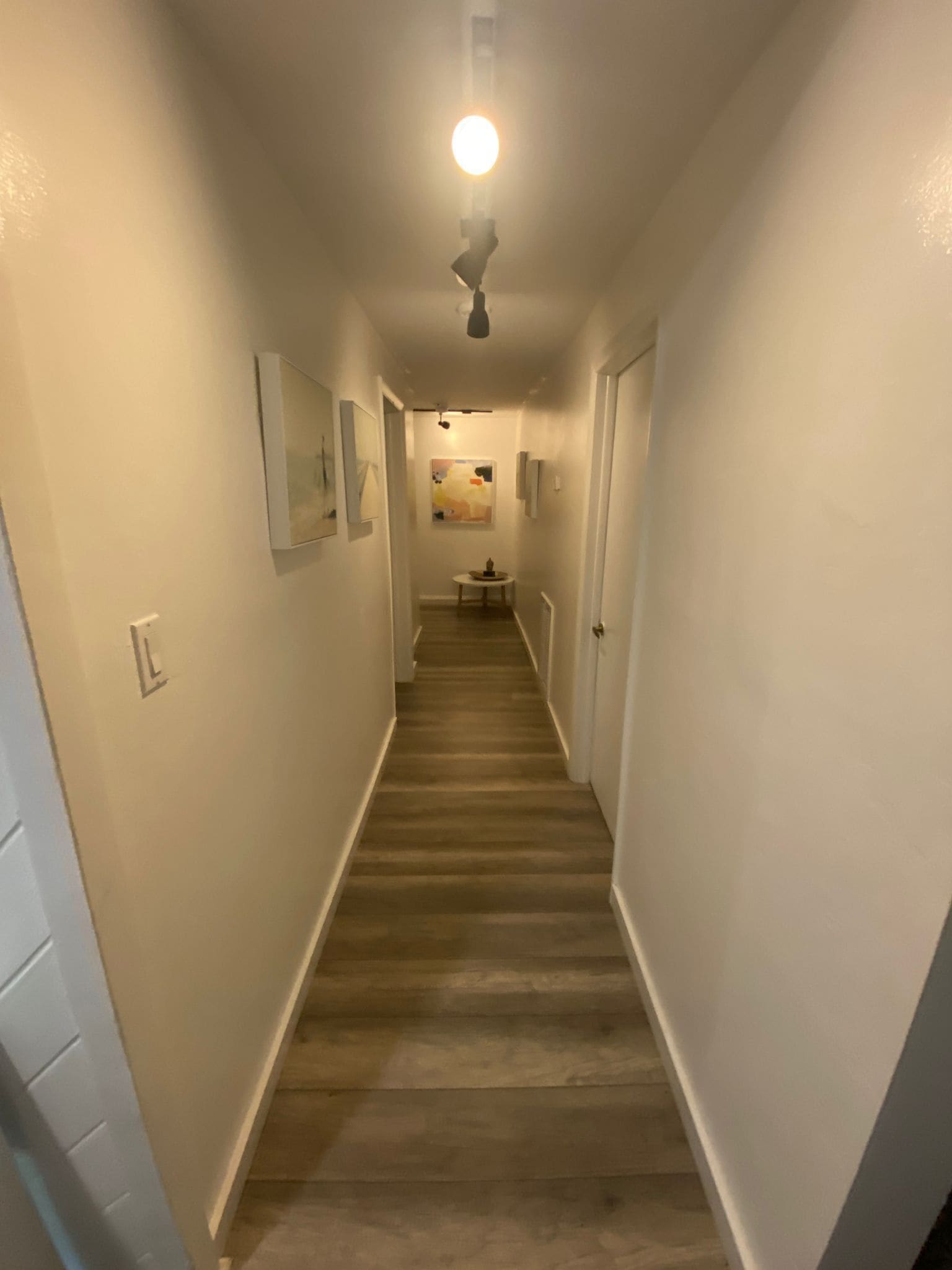 A narrow hallway with a wooden floor and white walls, leading to a door at the end.