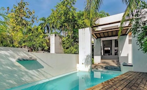 A swimming pool with a wooden deck, surrounded by lush greenery and a white building with a covered entrance.
