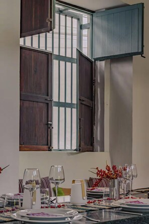 A dining area with a table set for a meal, a window with wooden shutters, and a view of the outside.