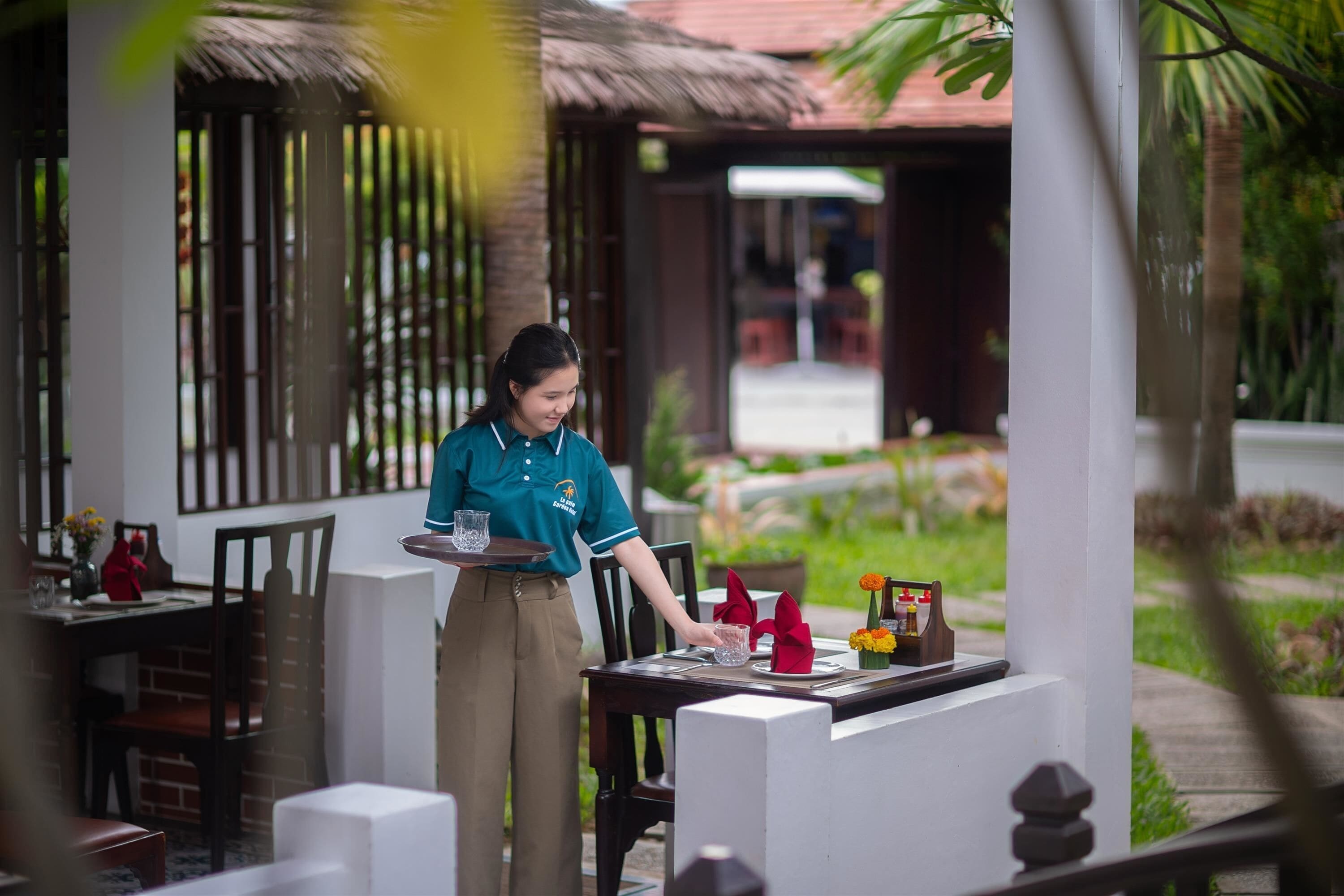 A waiter serving a table in an outdoor dining area with a thatched roof and greenery in the background.