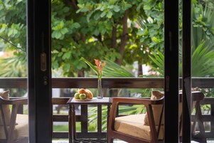 Um banco de madeira com almofada, uma mesa pequena com um vaso e frutas, e vista para o verde através de uma porta de vidro.