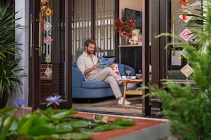 A person sitting on a blue sofa reading a magazine in a room with a glass door, a small table with a glass, and a vase with flowers.