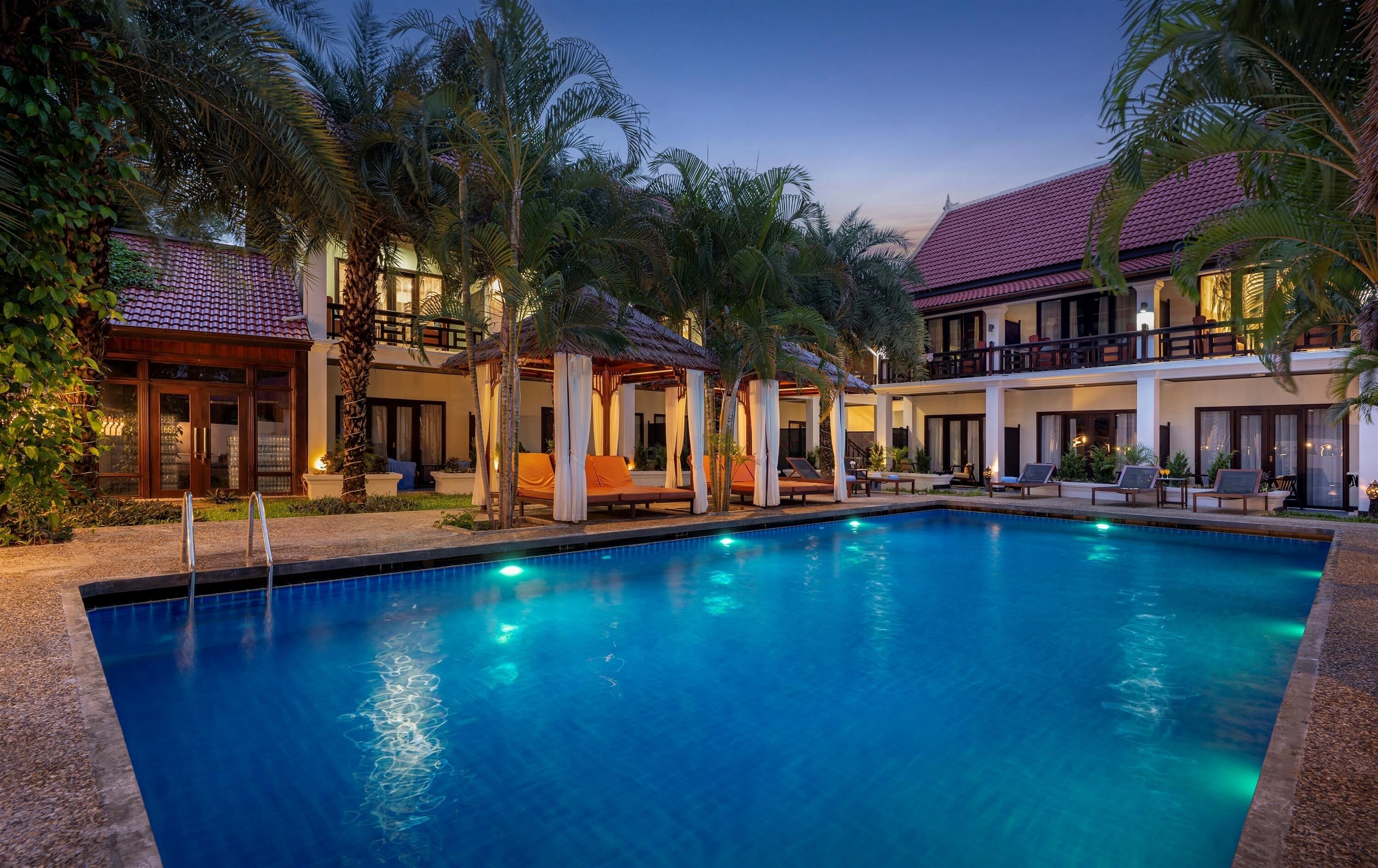 A swimming pool surrounded by lounge chairs and umbrellas, with a building featuring balconies and a tiled roof.