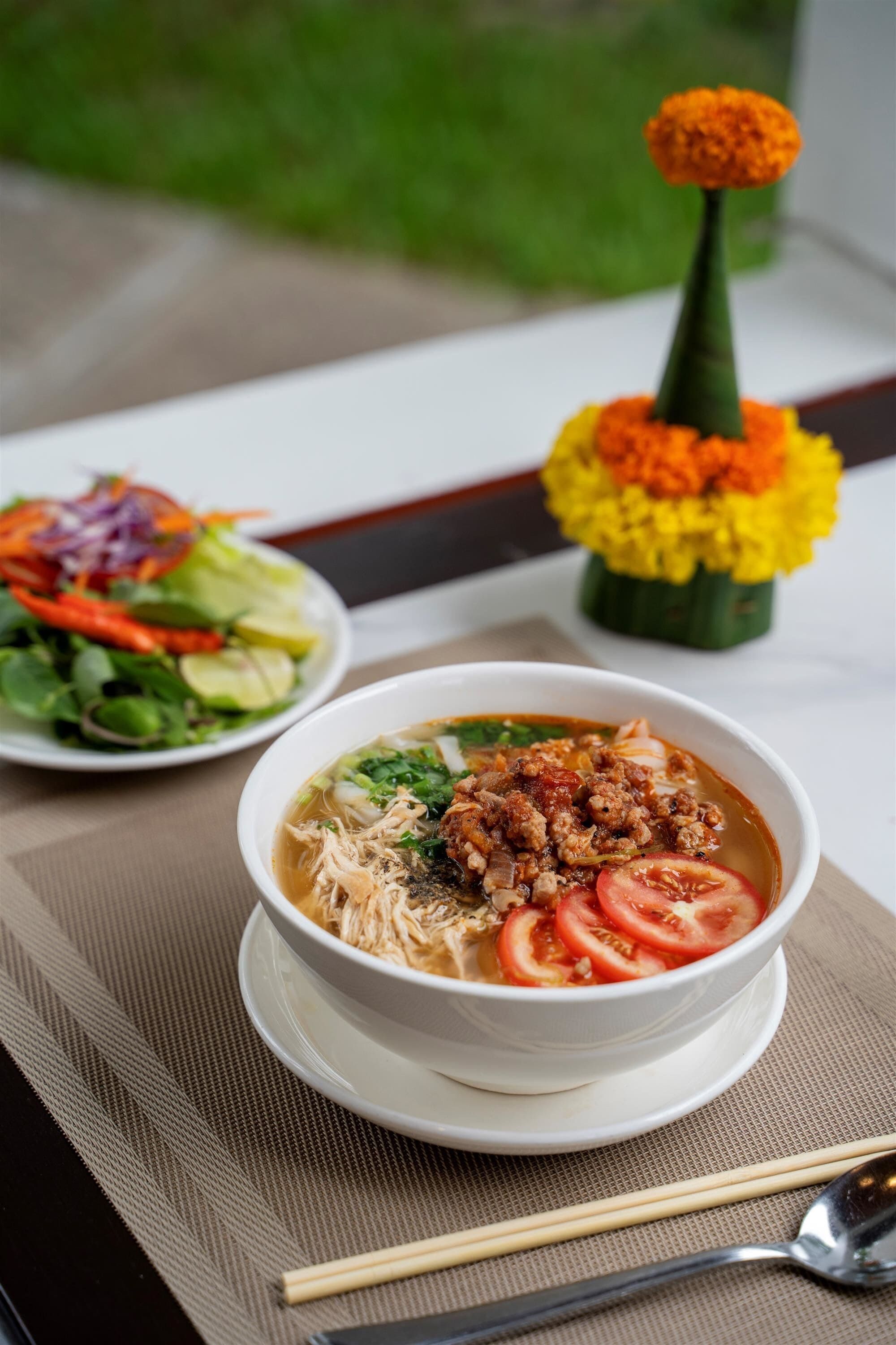 A bowl of soup with meat and vegetables on a table with a placemat, a vase of flowers, and a plate of salad.