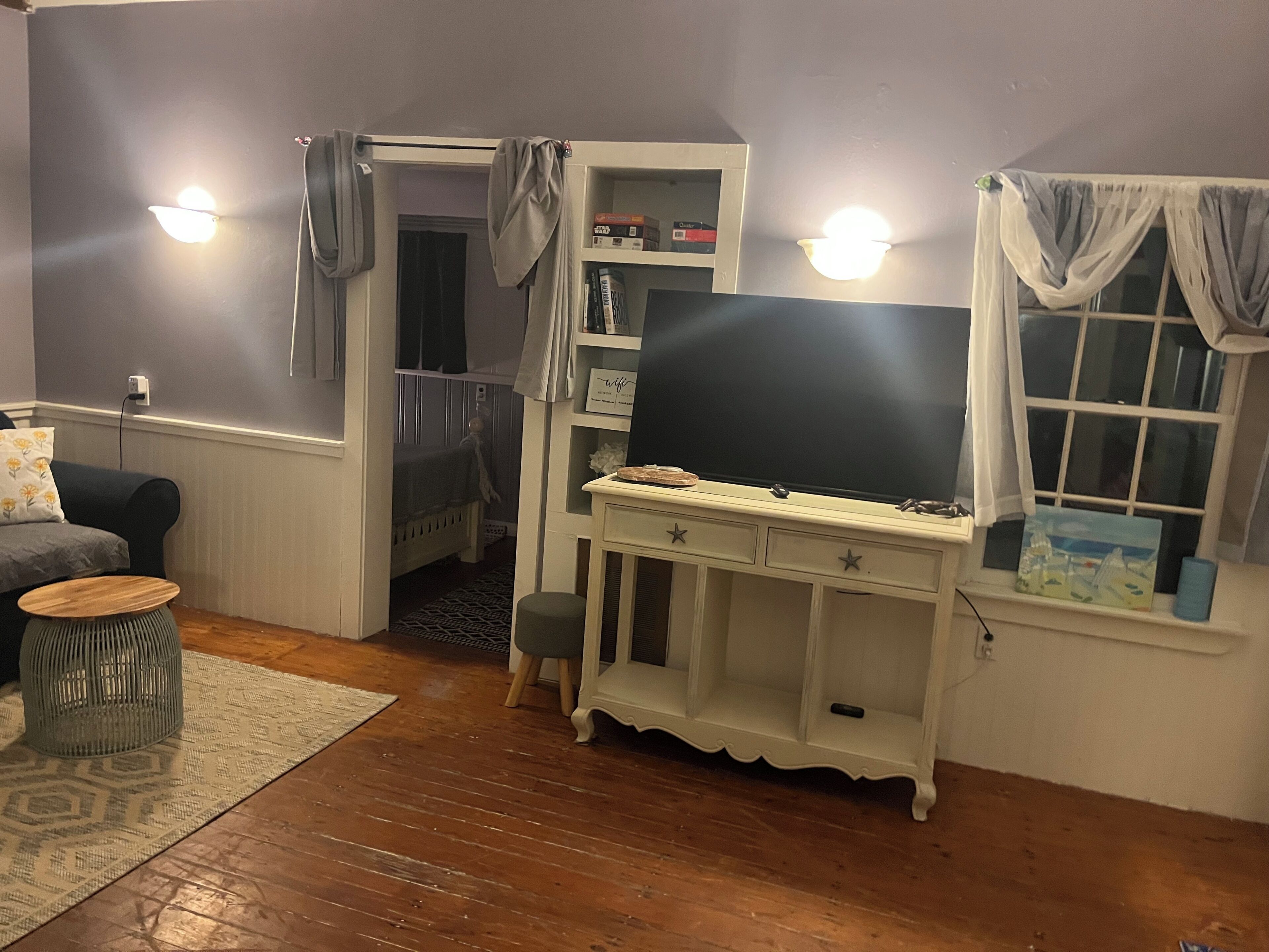 A room with a white desk, a television, a bookshelf, a window with curtains, and a door leading to another room.