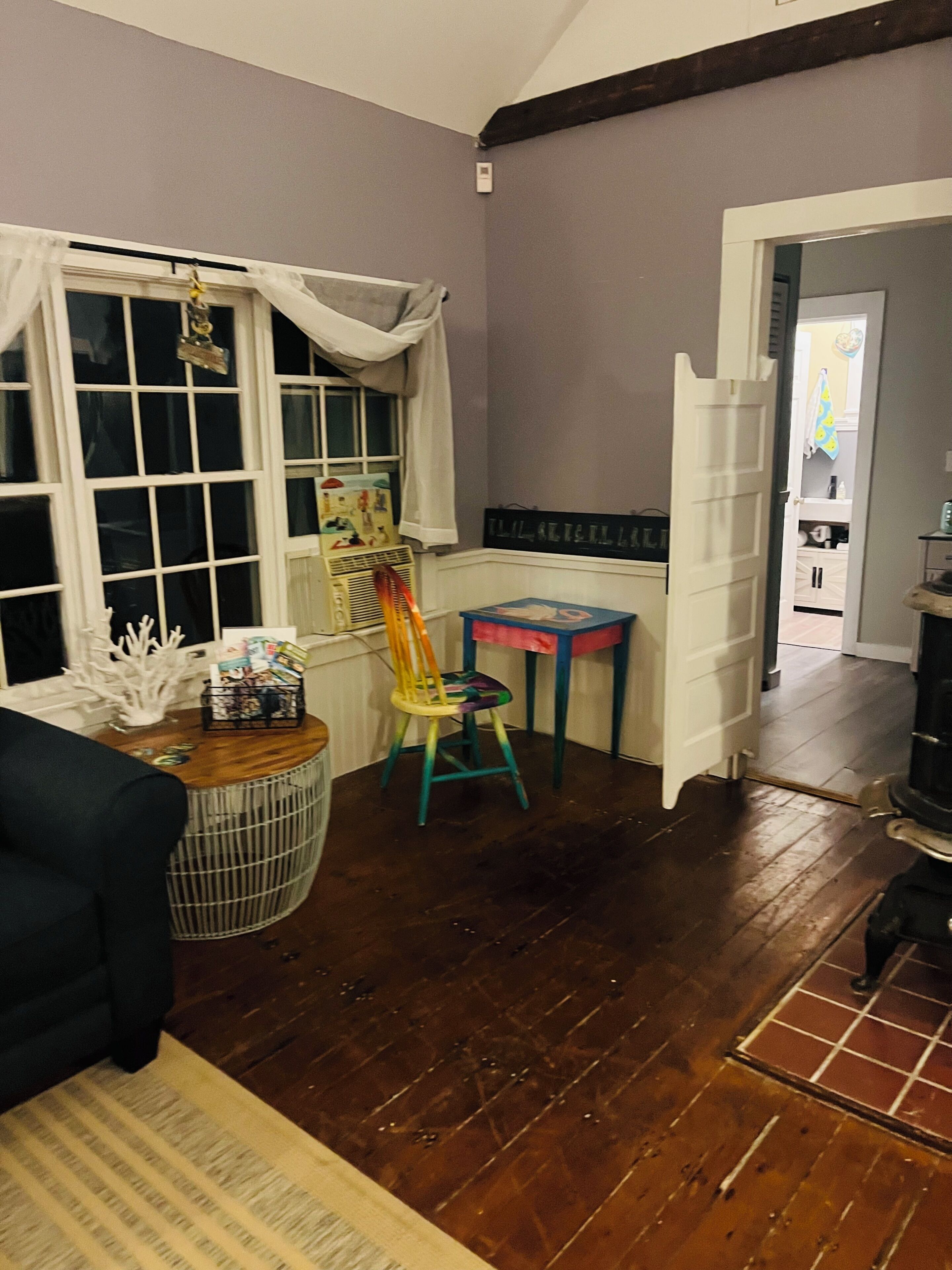 A room with a wooden floor, a window with curtains, a chair, a table with a basket, and a door leading to another room.
