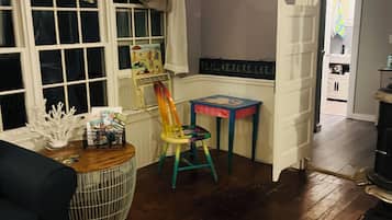 A room with a wooden floor, a window with curtains, a chair, a table with a basket, and a door leading to another room.