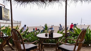 Ein Sitzbereich im Freien mit Korbstühlen und einem Tisch, mit Blick auf einen Strand, auf dem sich Menschen befinden.