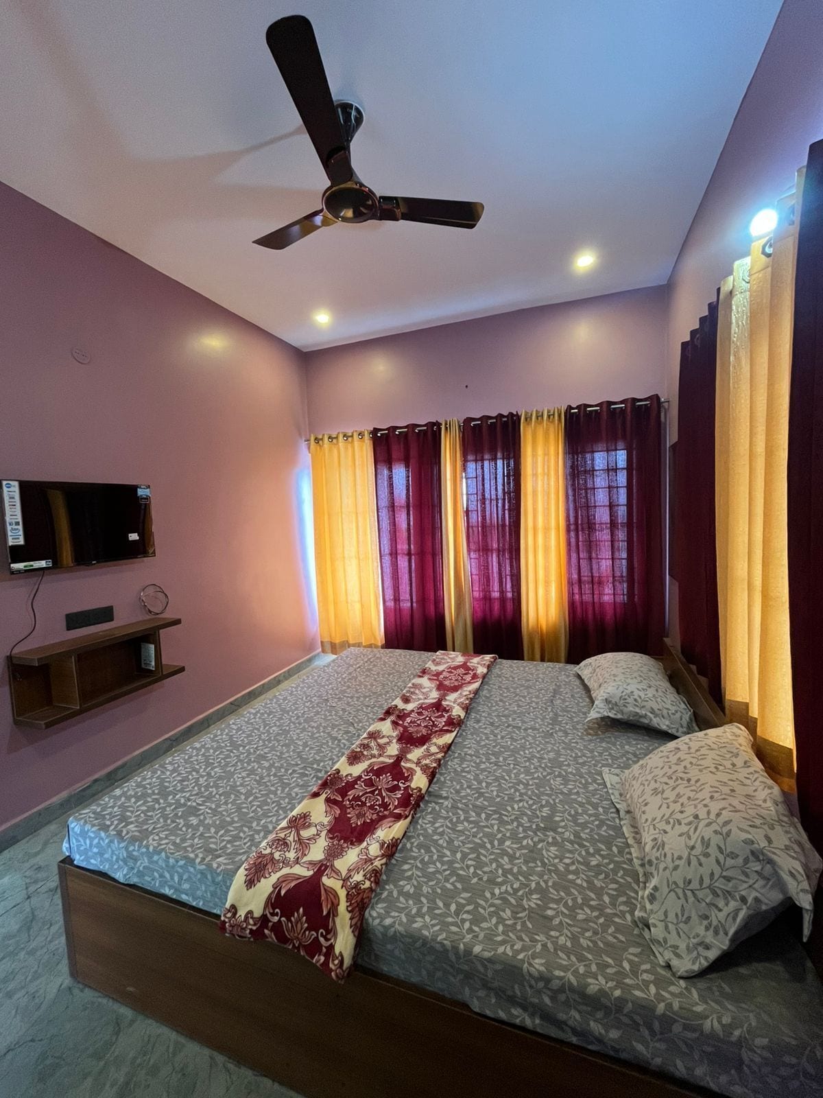 A bedroom with a bed, curtains, a ceiling fan, and a television on the wall.