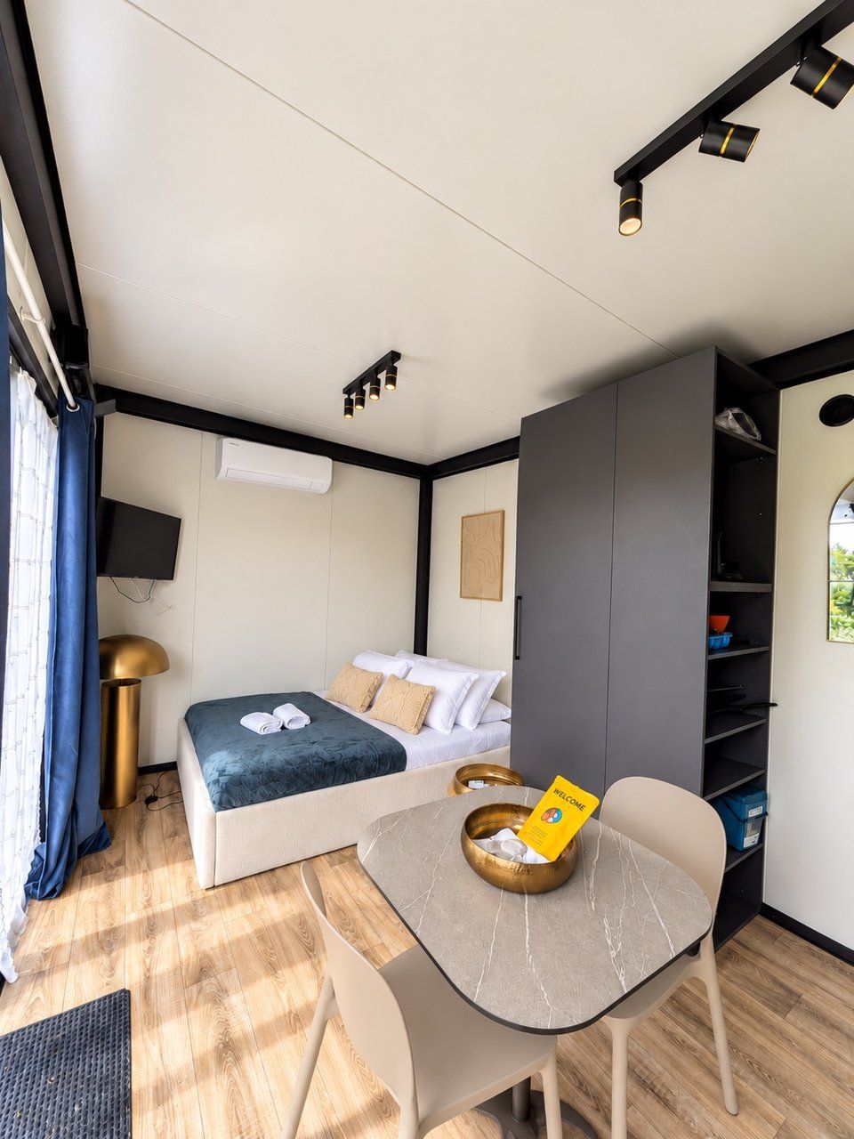 Quarto compacto com uma cama, uma mesa com um cartão de boas-vindas, televisão e guarda-roupa.