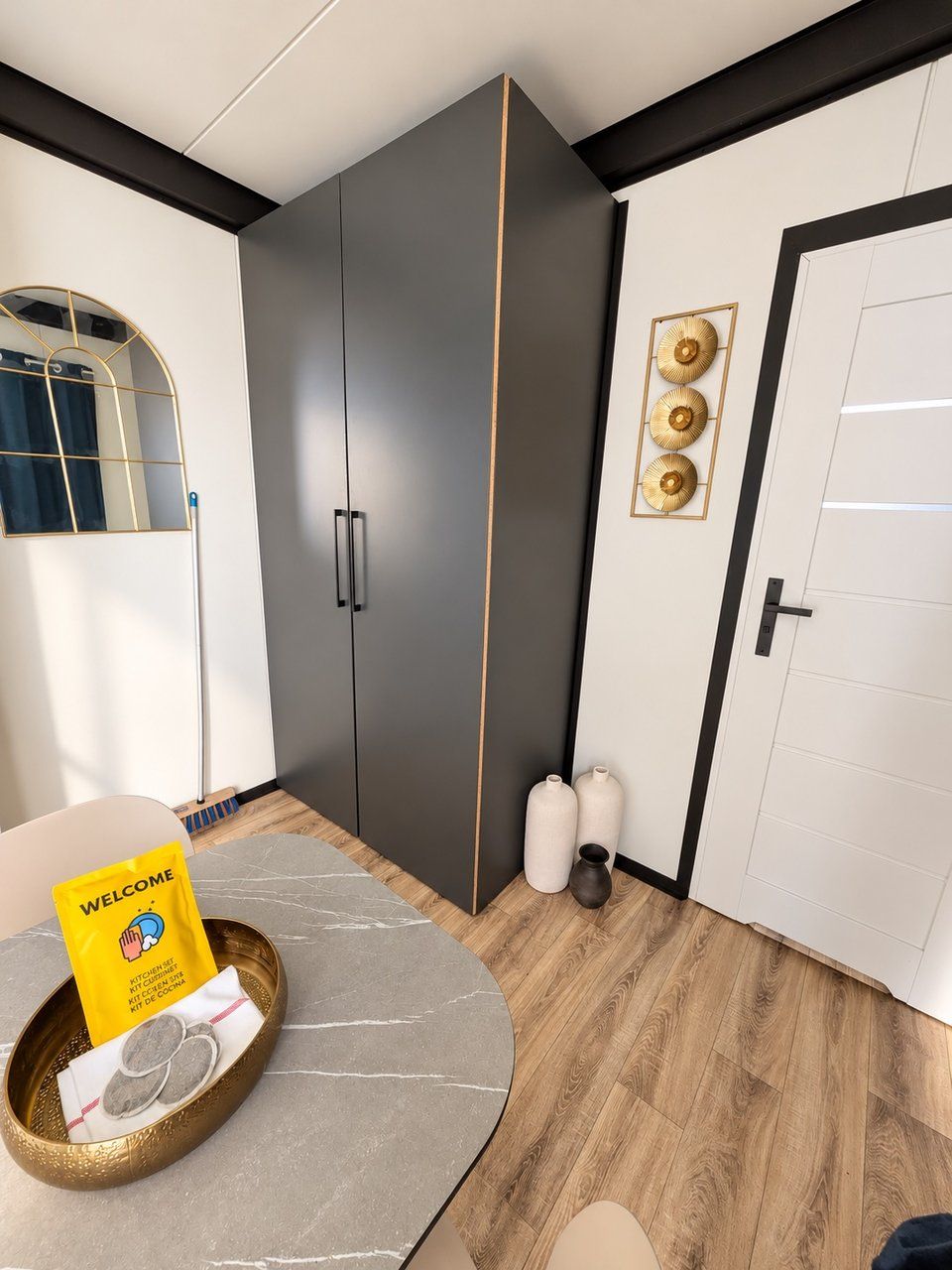 A modern room with a grey wardrobe, a round table with a welcome packet, and a door with a decorative plate.