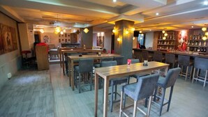 A restaurant interior with multiple dining tables and chairs, a bar area with shelves of bottles, and pendant lighting.