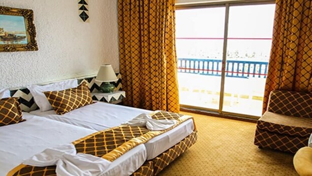 A hotel room with a bed, a chair, a window with curtains, and a view of a balcony.