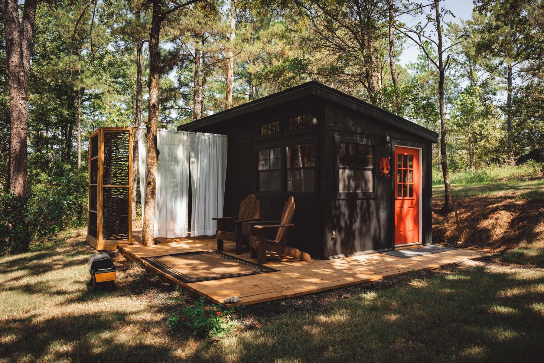 A small wooden cabin with a porch, a red door, and a grill outside.