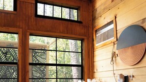 A wooden cabin interior with a bed, a window with a view of trees, a wall-mounted heater, and a decorative wall piece.