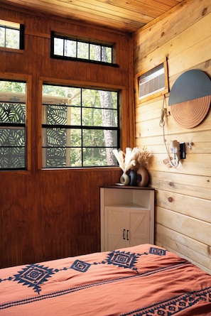 A wooden cabin interior with a bed, a window with a view of trees, a wall-mounted heater, and a decorative wall piece.