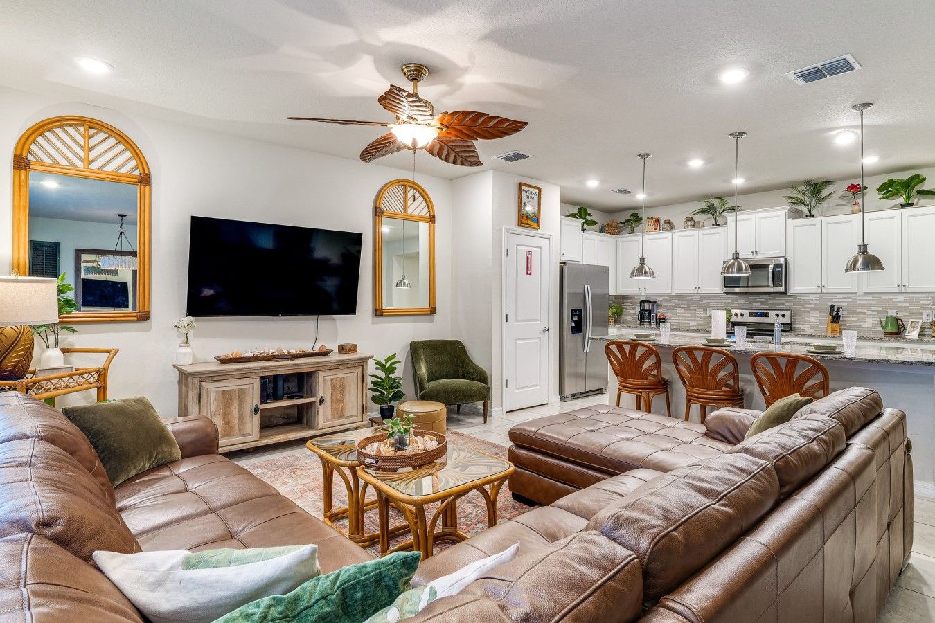 A modern living room with a large flat-screen TV, leather sofas, a dining area with wooden chairs, and a kitchen with white cabinets and stainless steel appliances.
