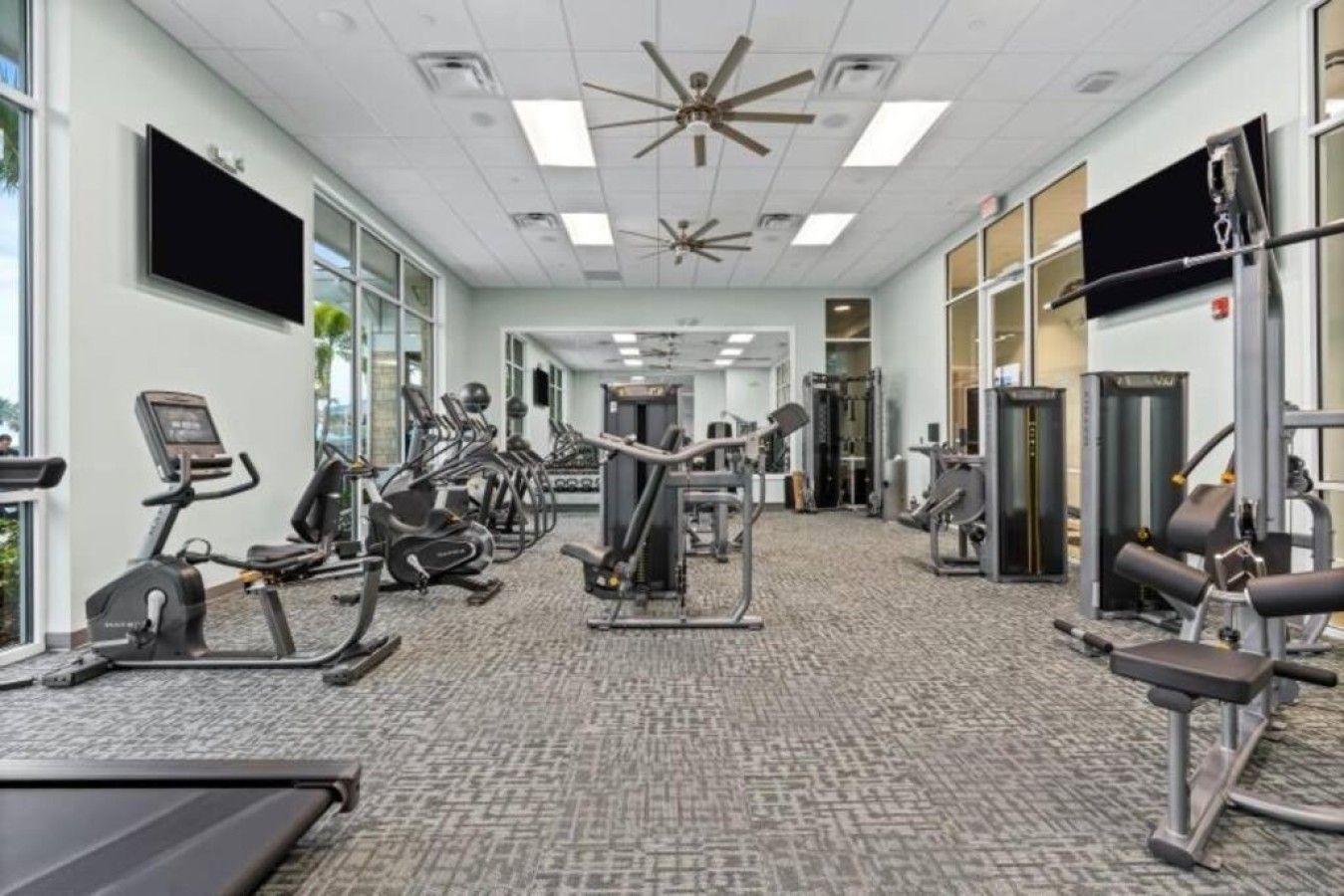 Un gimnasio con equipo de ejercicio variado, amplias ventanas y un ventilador de techo.
