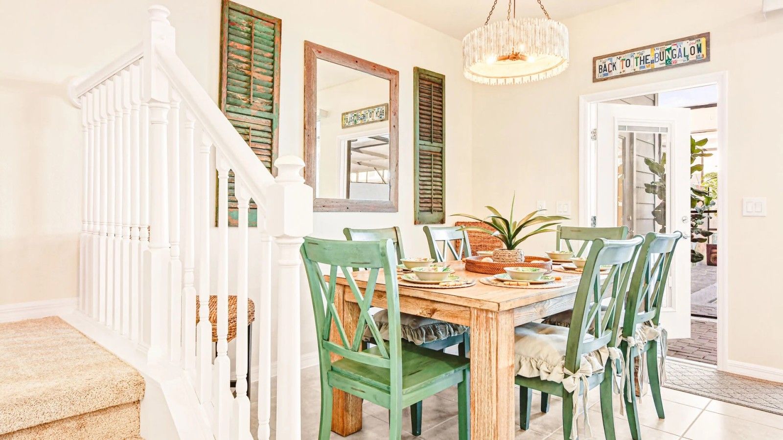 Uma sala de jantar com uma mesa de madeira, cadeiras verdes, um candelabro e uma placa com a inscrição 'VOLTA AO BANGALÔ'.