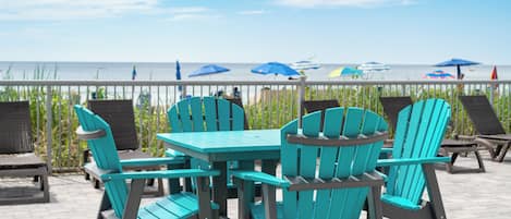 A patio with turquoise Adirondack chairs and a table, overlooking a beach with umbrellas.