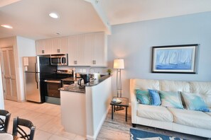 A modern kitchen with white cabinets, a black refrigerator, and a microwave. There is a dining area with a table and chairs, and a living room with a beige sofa and a painting on the wall. - Modern 2BR Oceanfront Condo w/ Balcony at Oceans One (Myrtle beach)