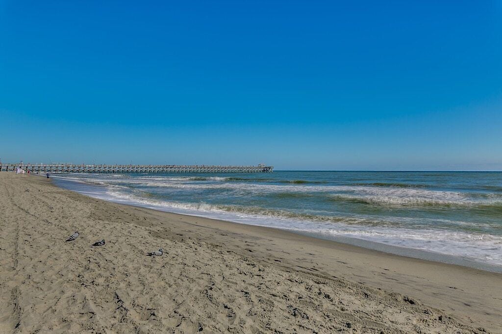A beach with a pier extending into the water, sandy shore, and a clear sky.