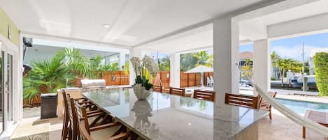 A spacious outdoor dining area with a large granite table, surrounded by wooden chairs, and a view of a swimming pool and tropical plants.