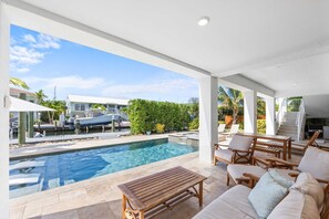 Zona de piscina con sillones de descanso y una mesa, un barco amarrado cerca y vistas de una zona residencial con casas y vegetación.