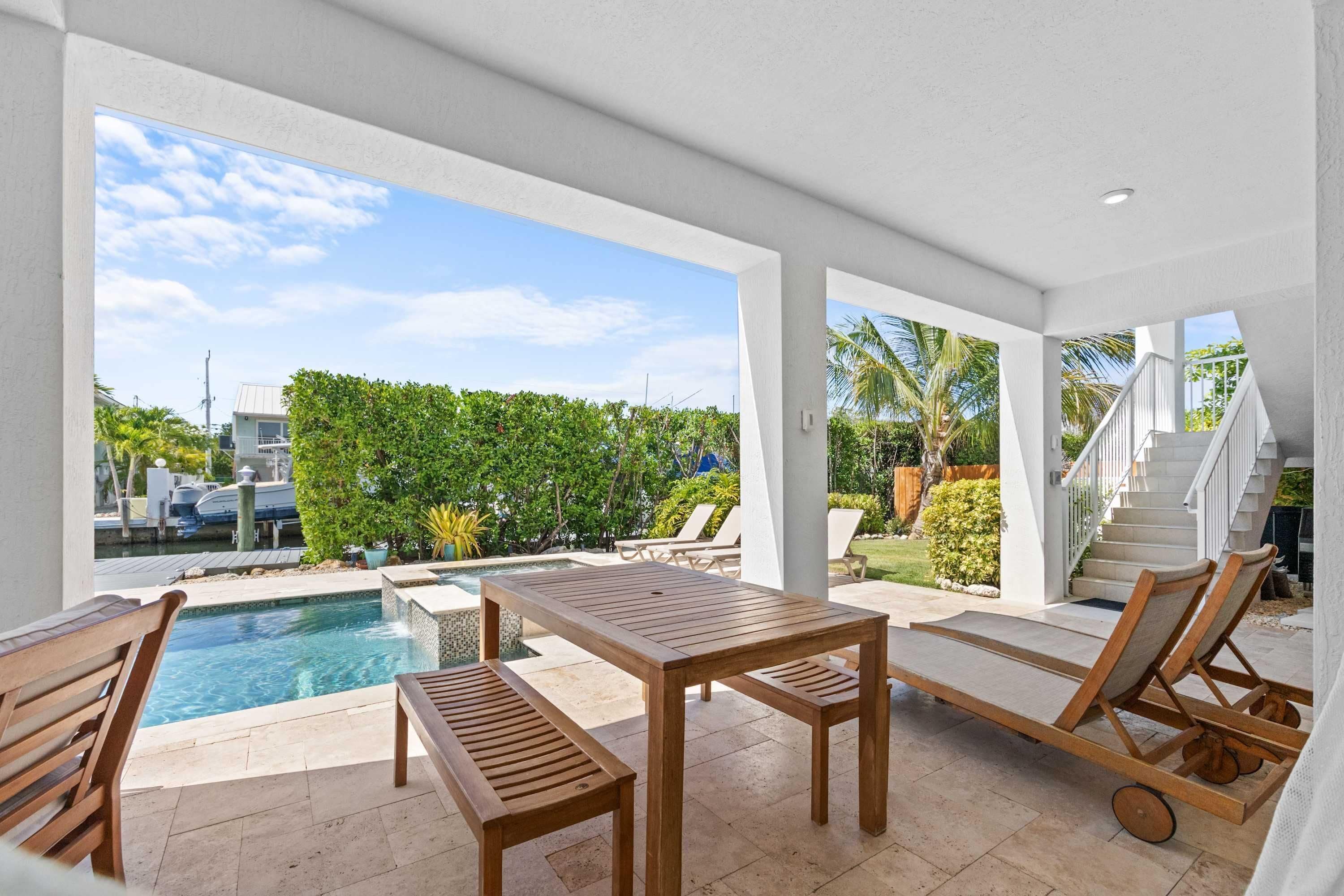 Uma área de piscina moderna ao ar livre com mesa de madeira e cadeiras de descanso, cercada por um muro branco e vista para uma paisagem tropical.