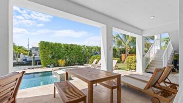 Uma área de piscina moderna ao ar livre com mesa de madeira e cadeiras de descanso, cercada por um muro branco e vista para uma paisagem tropical.