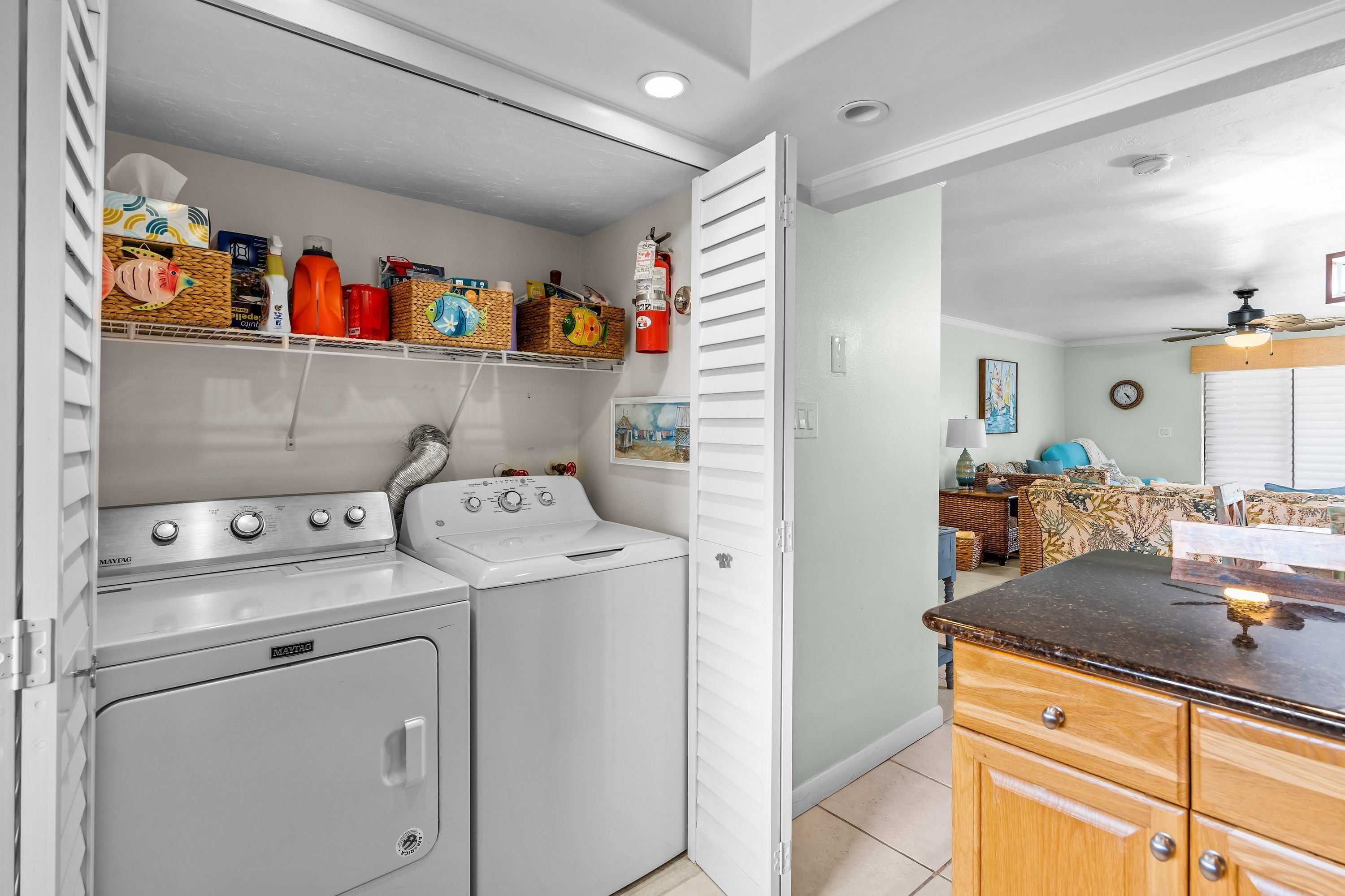 Un área de lavandería con lavadora y secadora Maytag, un estante con diversos objetos, y un comedor con mesa y sillas.
