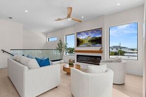 A modern living room with a large flat-screen TV, a ceiling fan, and a view of the outdoors through large windows. - Cielo del Mar - Waterfront - Heated Pool - Dock (Marathon)