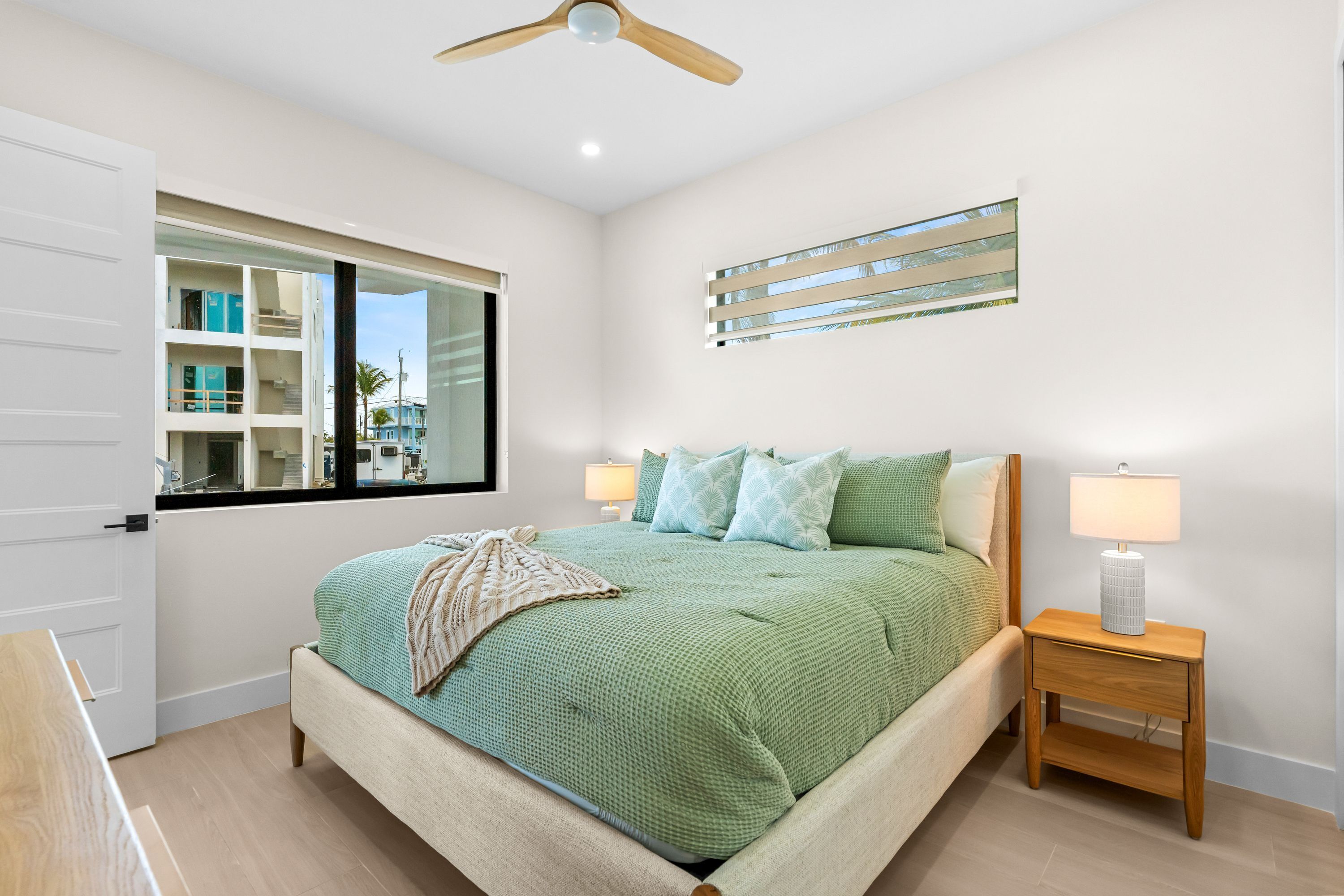 A bedroom with a bed, two bedside tables with lamps, a window with a view, and a ceiling fan.
