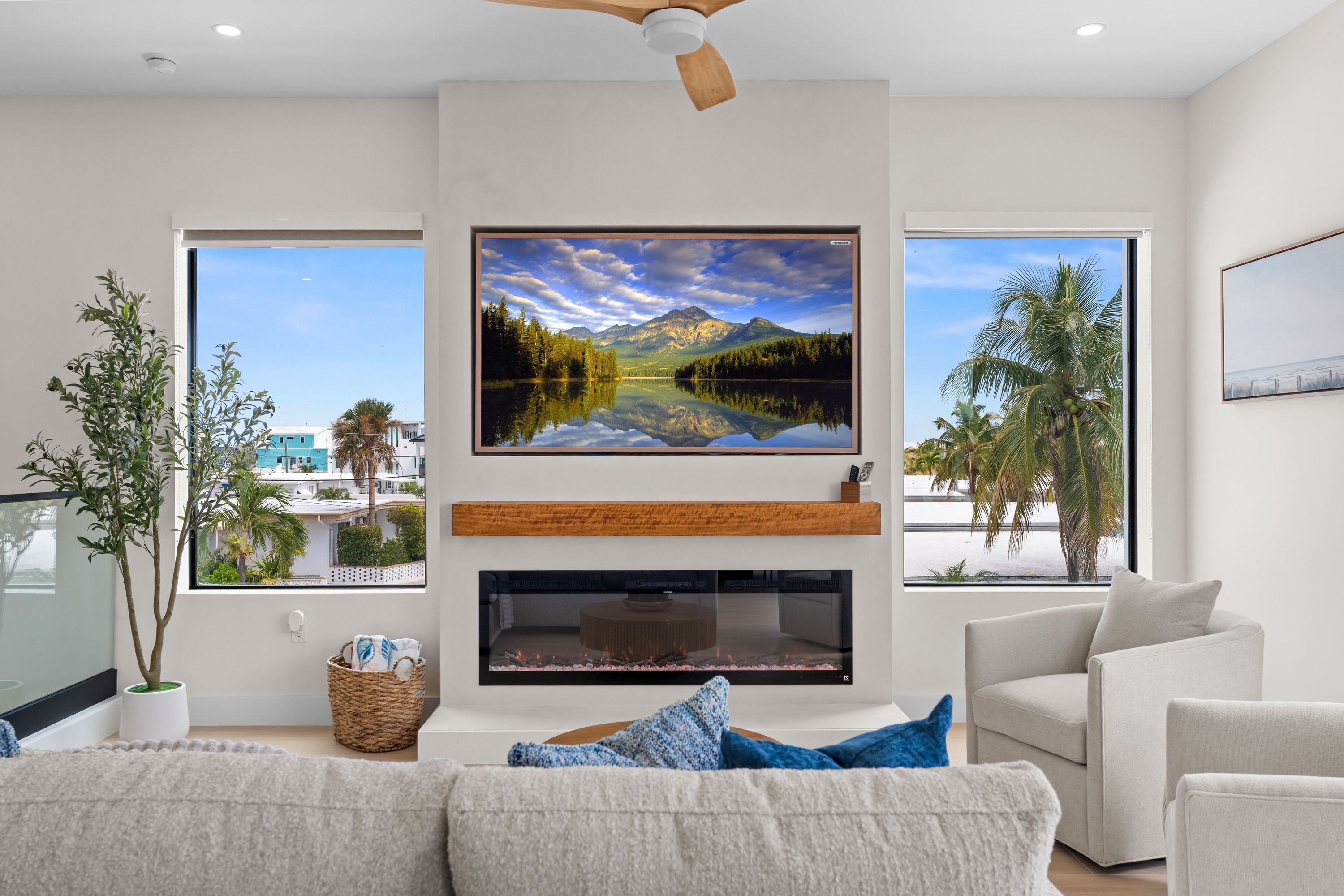 A modern living room with a fireplace, a large TV mounted on the wall, and two windows offering views of palm trees and a lake.