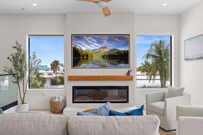 A modern living room with a fireplace, a large TV mounted on the wall, and two windows offering views of palm trees and a lake. - Cielo del Mar - Waterfront - Heated Pool - Dock (Marathon)