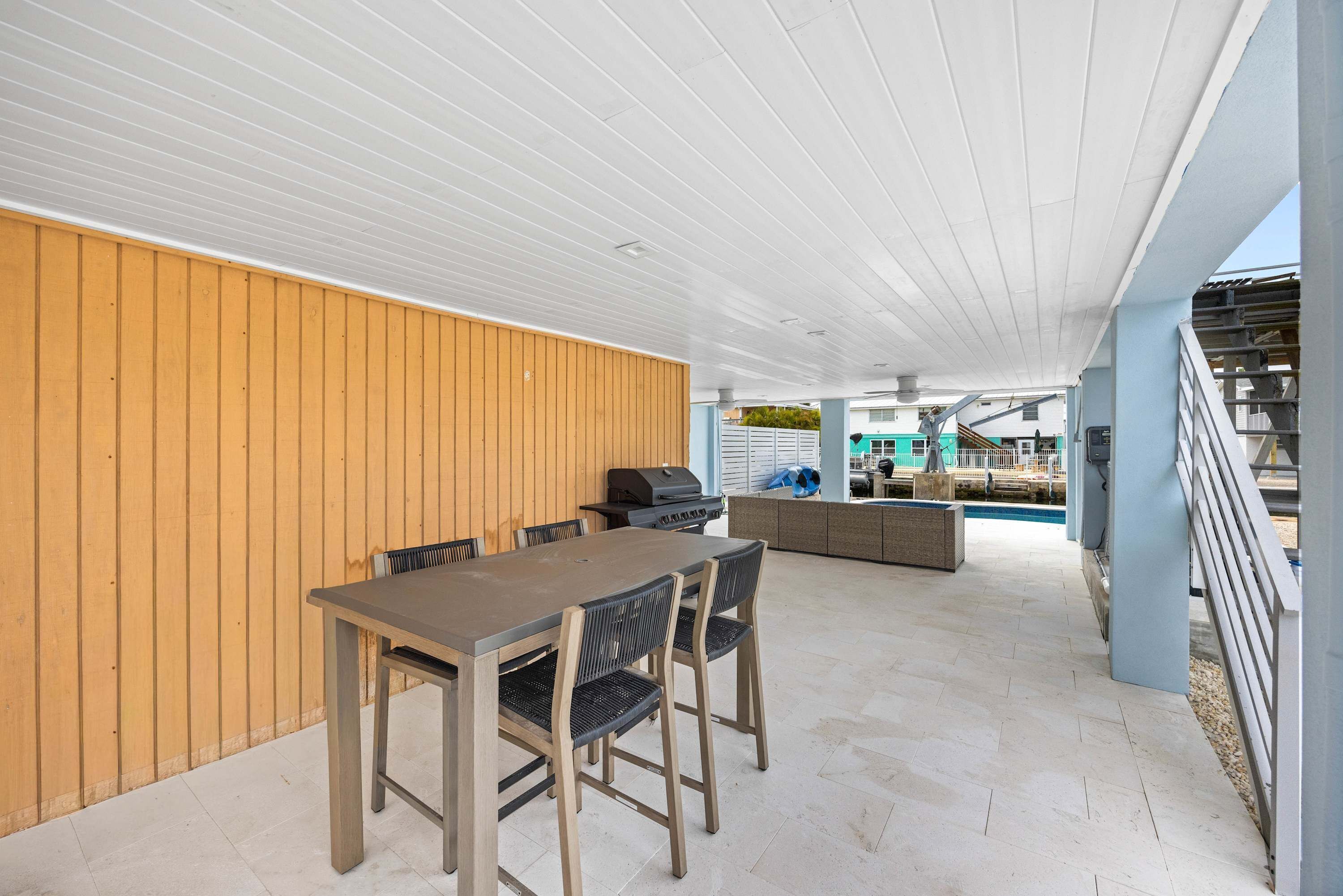 An outdoor patio area with a table and chairs, a grill, and a view of a swimming pool and buildings in the background.