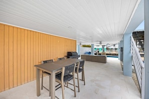 An outdoor patio area with a table and chairs, a grill, and a view of a swimming pool and buildings in the background. - Catch of the Bay - Waterfront - Pool - 35 Dock (Marathon)