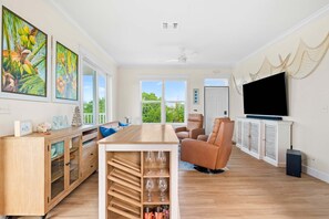 A spacious living room with a wooden dining table, comfortable seating, a flat-screen TV, and large windows offering a view of greenery. - The Salty Coconut Waterfront Dock Hot Tub (Marathon)