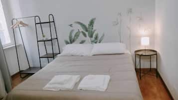 Un letto rifatto con due asciugamani bianchi piegati, una mensola in metallo nero, una pianta sospesa al muro e un comodino con una lampada.