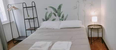 Una cama bien hecha con dos toallas blancas dobladas, un mueble metálico negro, una planta colgante en la pared y una mesita de noche con una lámpara.